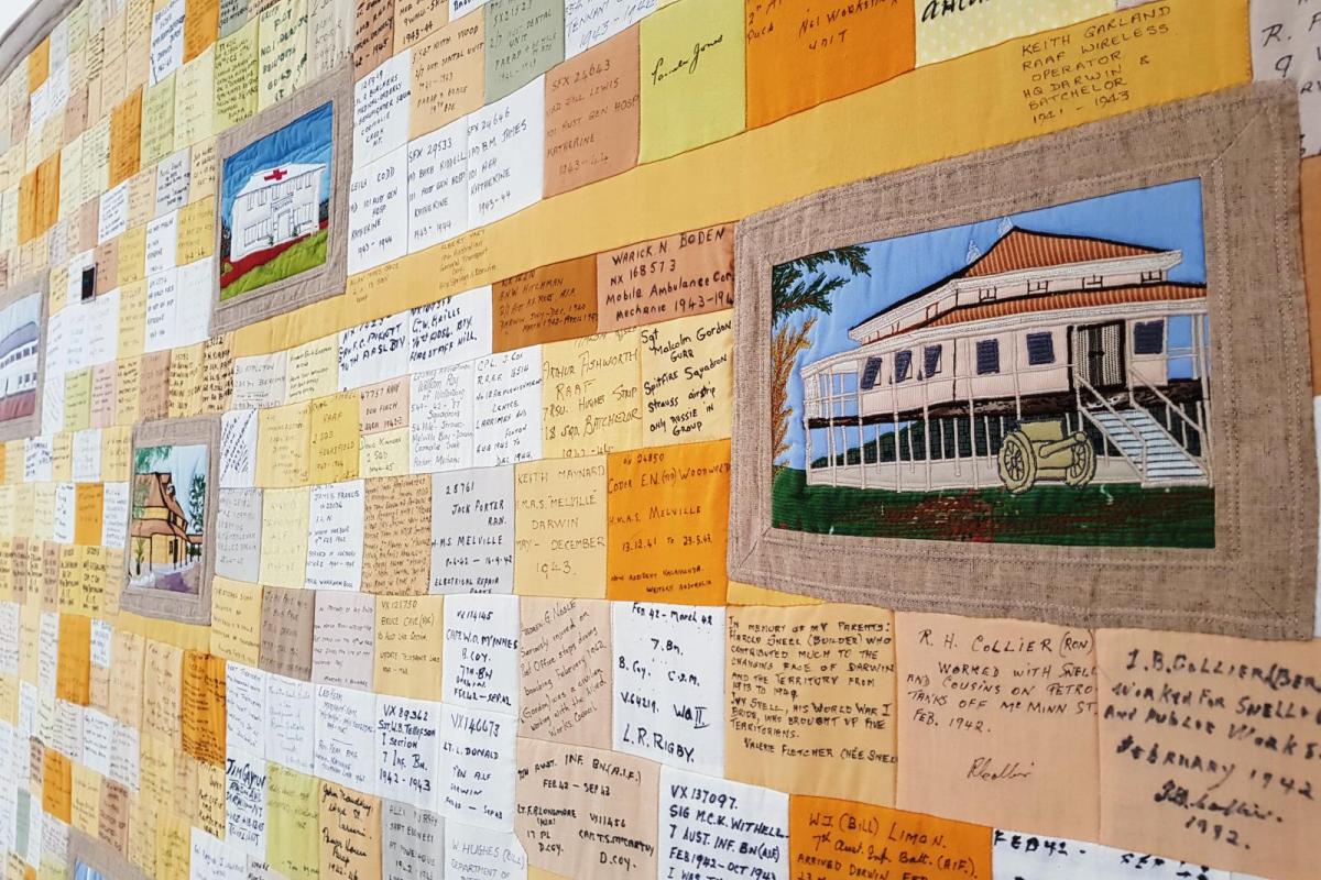 Section of the Commemorative quilt. Photo is angled across the quilt revealing oranges toned patches with black writing on patches. Larger patches of iconic Darwin images scattered throughout names. 