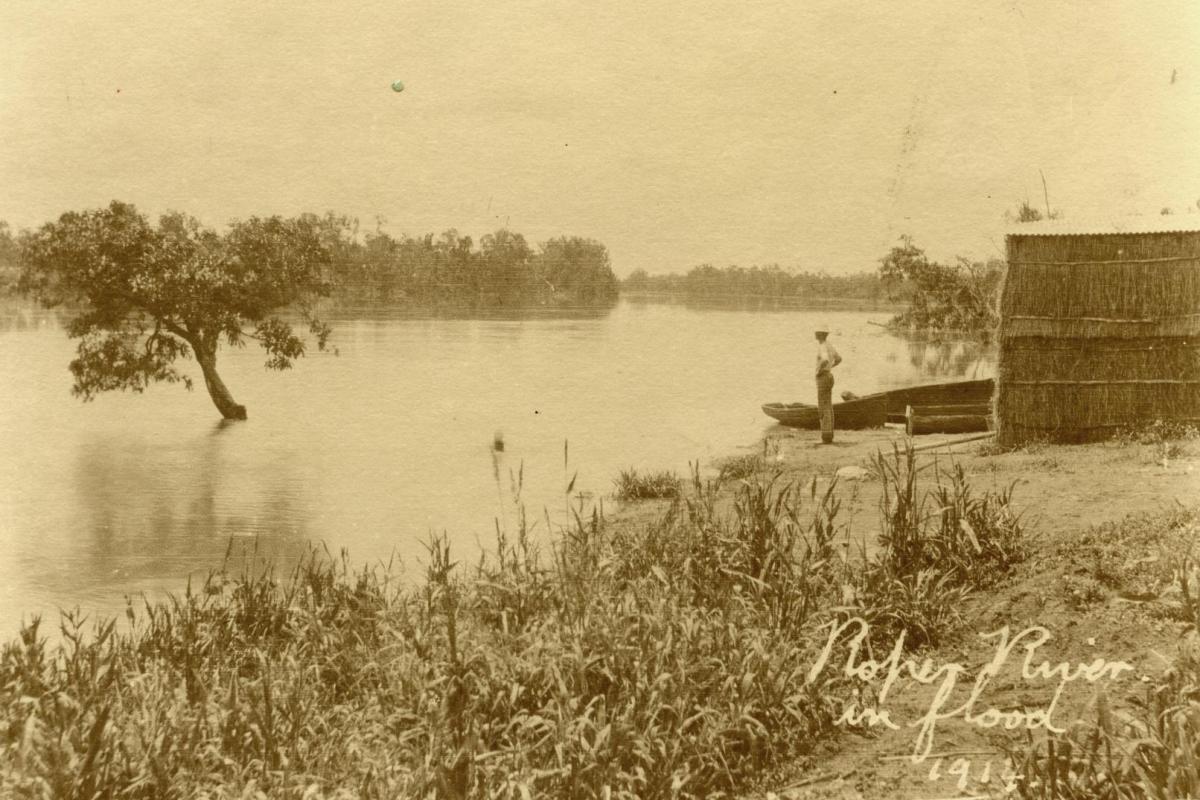 Sepia colour image of man standing on bank and looking at the Roper River with a stack of hay bales behind him