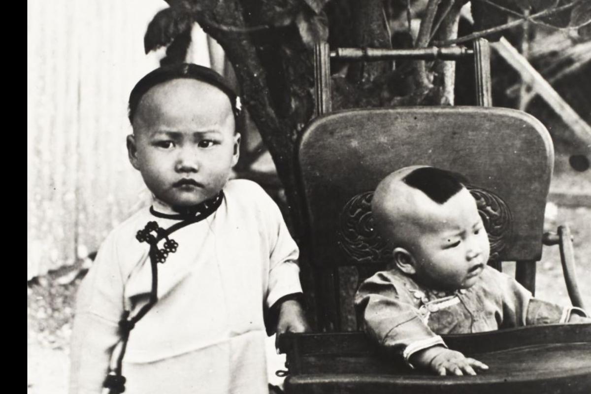 Black and white photo. A young child and a baby. The baby is seated in a wooden pushchair 
