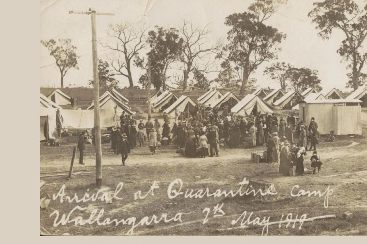 Large group of men and women arrive at camp of tents, with toilet block at right, power pole at left and gum trees in the distance.