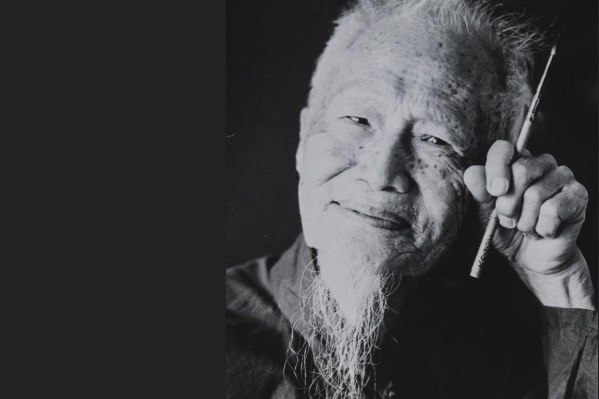 Black and white photo. An elderly man is smiling. His hand, which is held to his face grips a paintbrush between his index and forefinger 