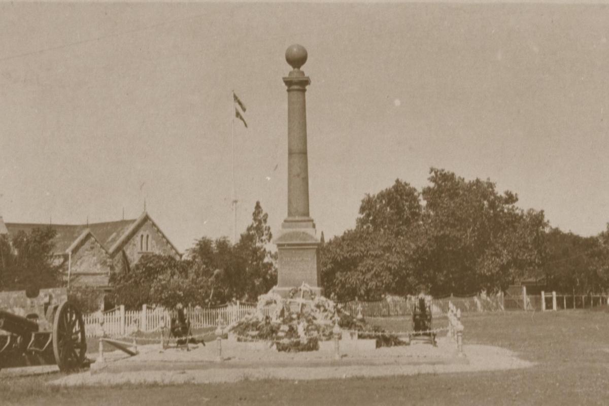 The Cenotaph stands at centre with floral wreaths at its base. In the background is Government House and two machine guns. At left is a field gun.
