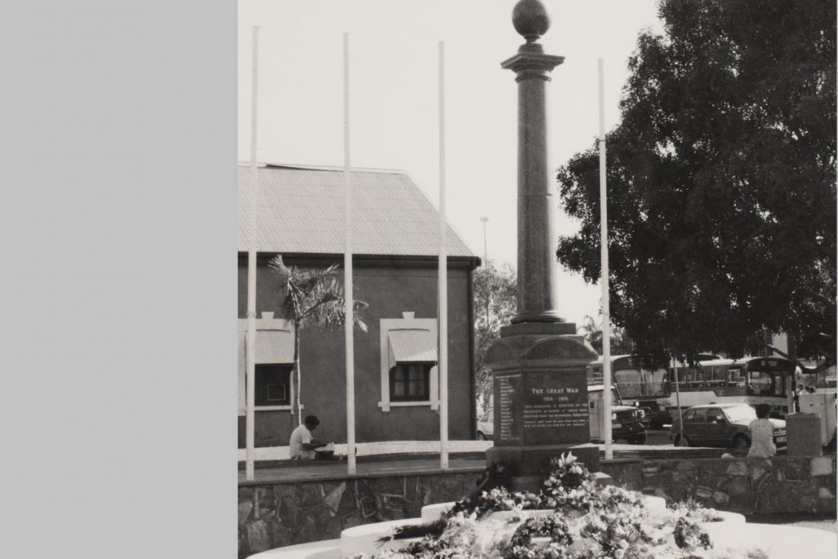 The Cenotaph is at centre with floral wreaths at its base. In the background at left is a building (Browns Mart) and at right is a large tree. A bus can be seen in the far distance.