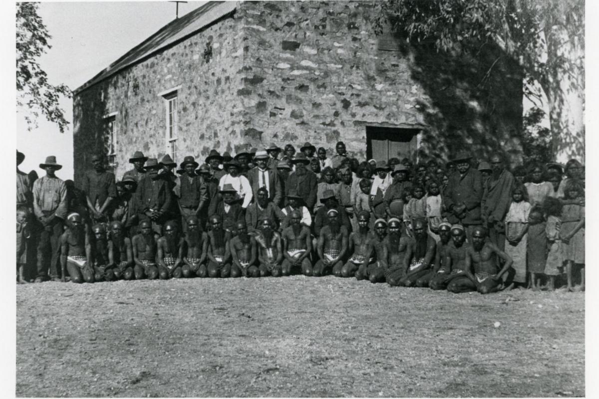 Depicts a large group of Aboriginal people in front of a large stone building.