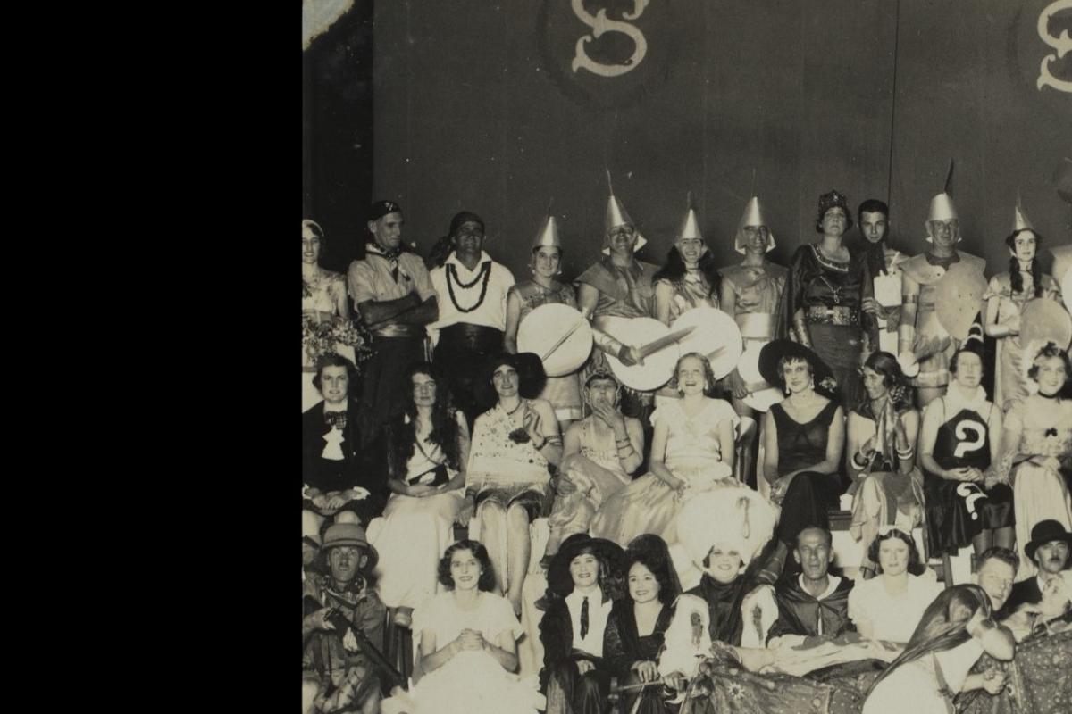 Among a large gathering of people seated in fancy dress are three men dressed in women’s clothing.