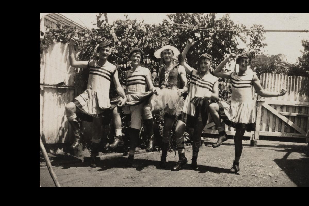 Five men in a “revue” pose, wearing short dresses, corsets, hats, make-up and boots.