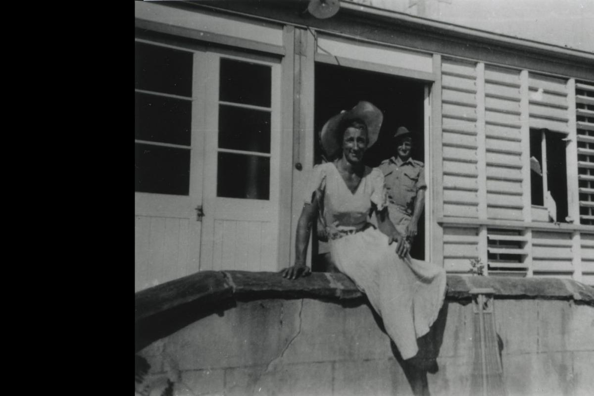 Smiling man wearing summer dress and hat sits in front of damaged building with a smiling soldier in doorway.