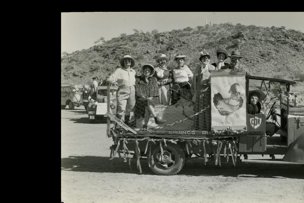 Seven masked people in women’s clothing standing on the back of a truck with a sign reading “Ye Olde Hens”.