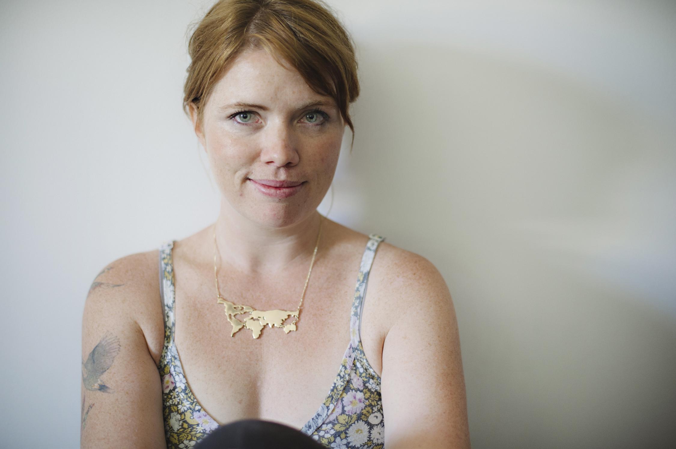 Woman with red hair and wearing a gold necklace and singlet in seated position posing for the camera