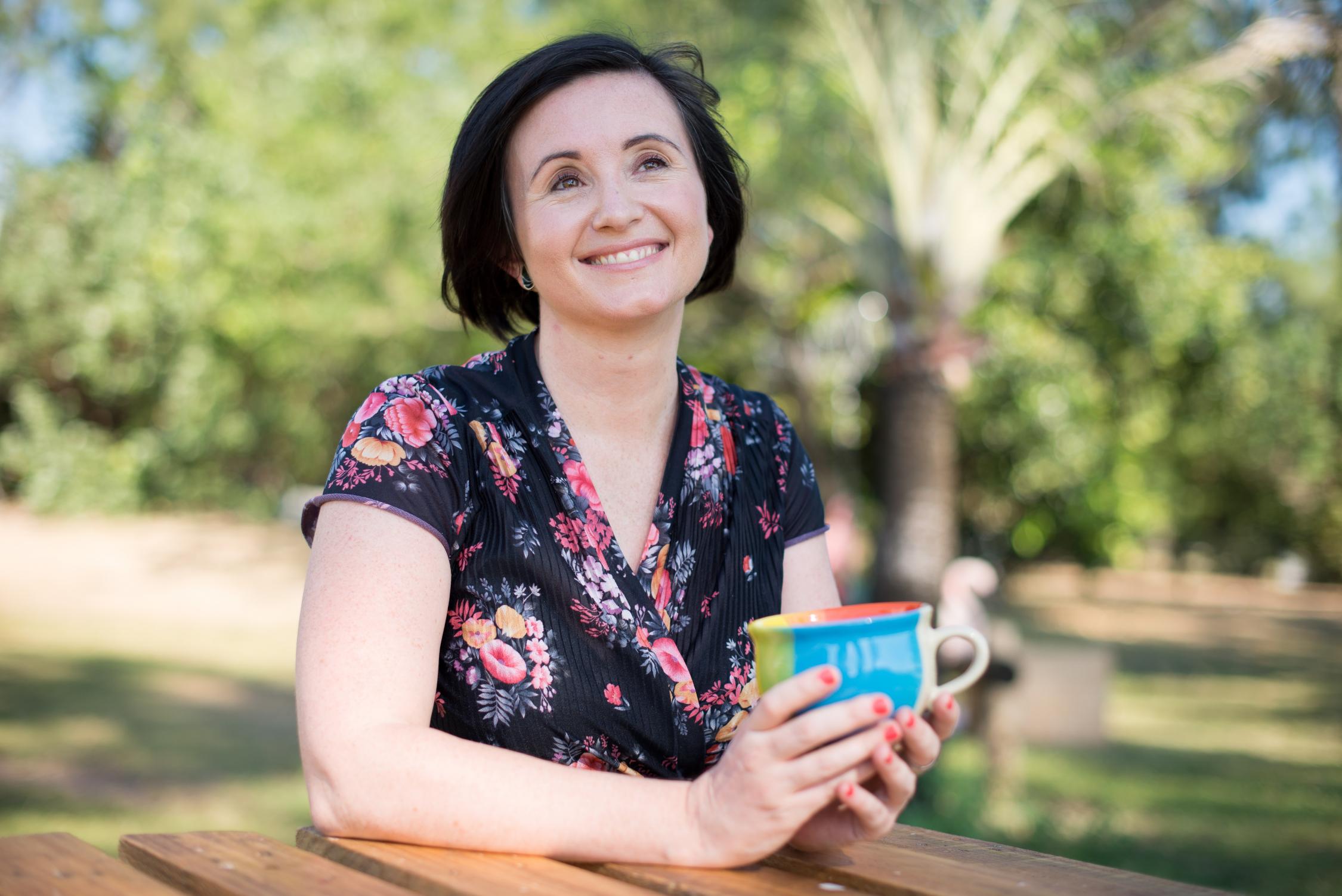 A woman with shot bobbed brown hair sitting at a bench outside holding a cup in her hands and gazing skywards