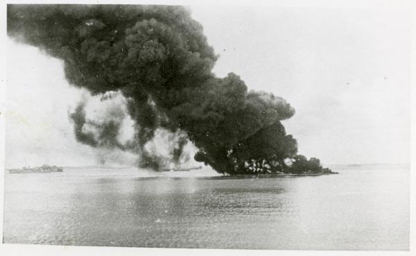 The impact of the first air raid on shipping with (L-R) the HMAS Swan; Floating Dock AFD18; SS Zealandia burning and sinking; HMAS Wato (tug); HMAS Platypus (obscured); oil burning from sunken USS Peary.