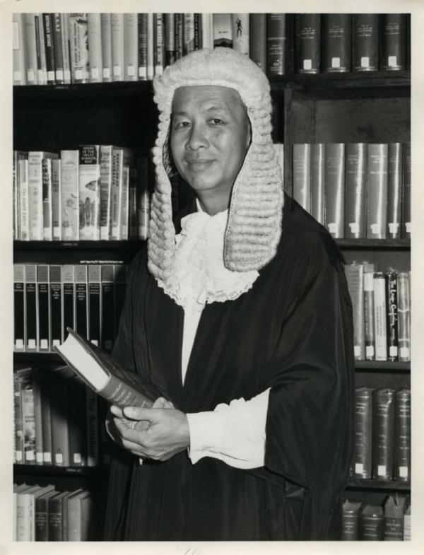 Black and white photo. Man in court wig and gown standing in front of a bookcase and holding a book.