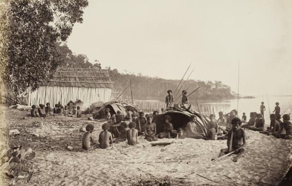 Depicts a group of Aboriginal men, women and children on the beach with traditional wurlies in the foreground and a European style building in the background.