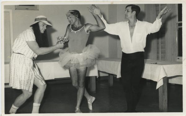 Two male dancers, one in a leotard and tutu, pose with an actor in drag who is handing them a bouquet of flowers. 