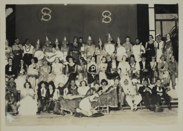 Among a large gathering of people seated in fancy dress are three men dressed in women’s clothing.