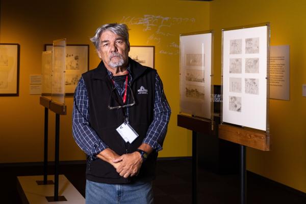 Man standing in front of framed drawings, displayed in a dimly lit room. 